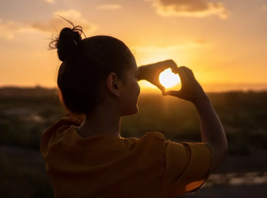 Photo d'une femme qui fait un cœur avec les mains face à un levé de soleil. Respect de soi.