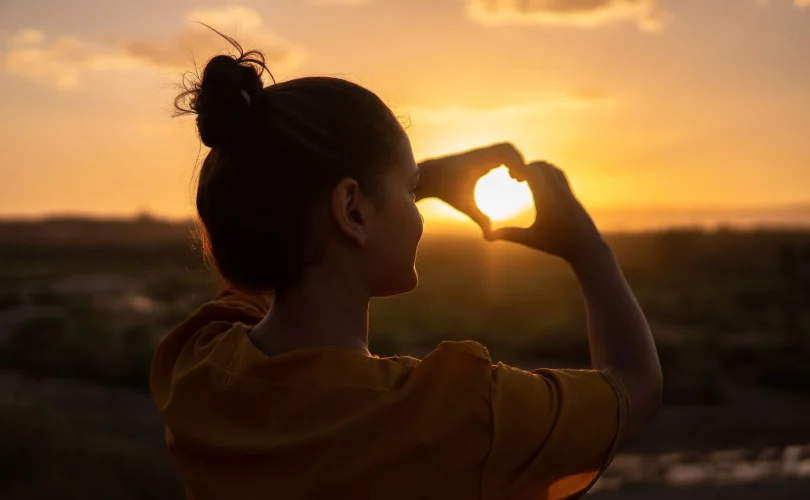 Photo d'une femme qui fait un cœur avec les mains face à un levé de soleil. Respect de soi.