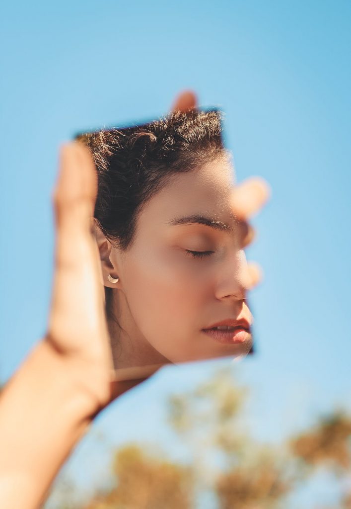 Une photo d'une femme brune les yeux fermées, qui est face à son reflet dans un miroir qu'elle tient à la main. C'est une photo qui représente le respect de soi.