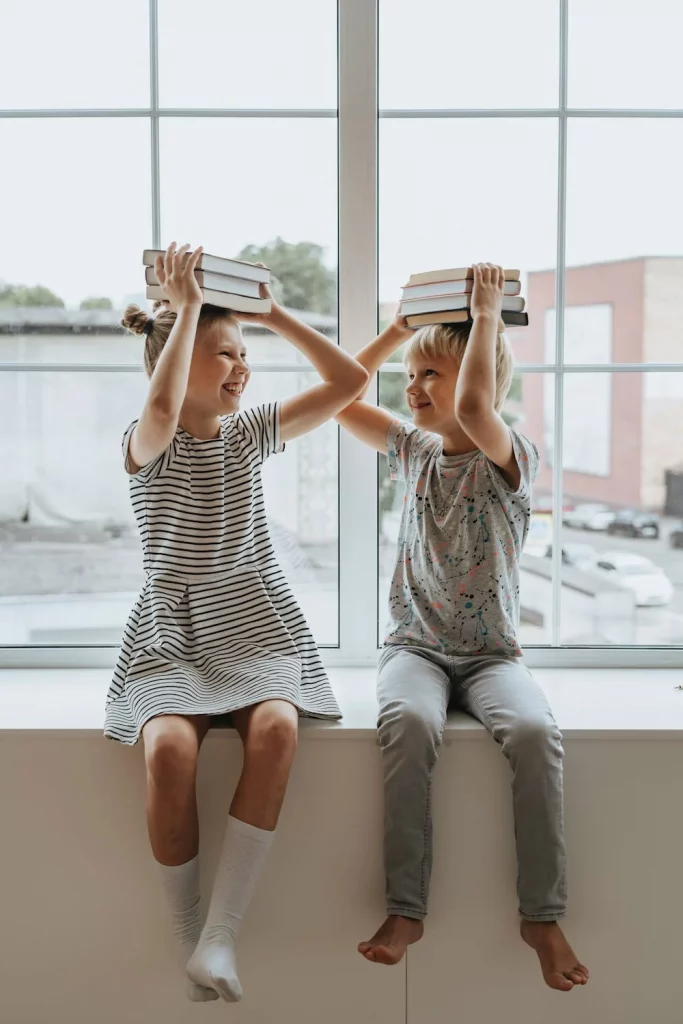 Photo de deux enfants, un garçon et une fille, assis sur le rebords d'une fenêtre à l'intérieur d'un bâtiment. Ils tiennent des livres sur leurs têtes et rient, en cours d'éducation sexuelle.